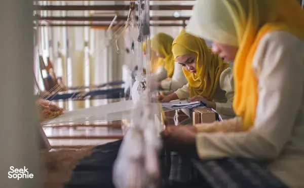 Weaver in a yellow headscarf crafting on a loom at Tanoti Crafts workshop in Kuching, Sarawak, with students working nearby