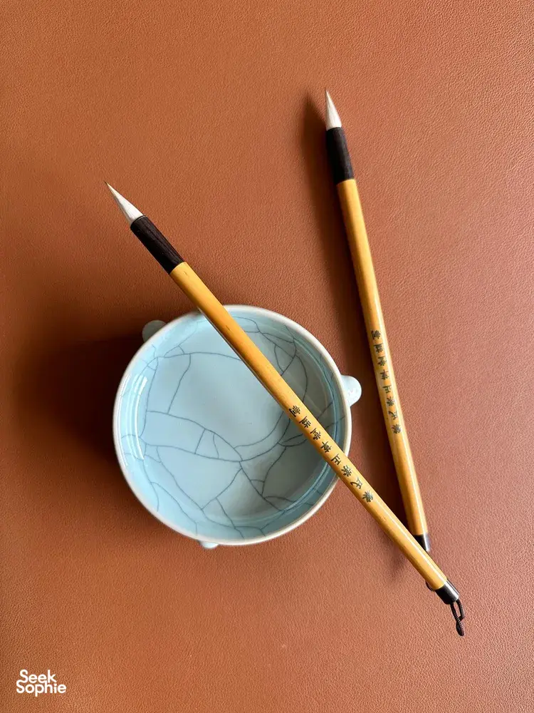 Two Chinese calligraphy brushes beside a small blue cup on a peach background, ready for beginner writing in Singapore—workshops in Singapore