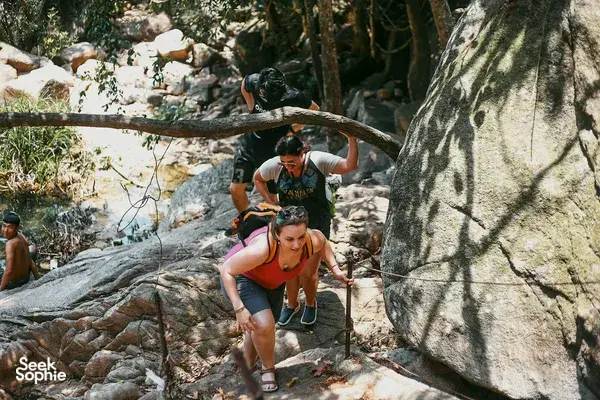 Hikers clambering over wet rocks using a rope line in a jungle stream during a Hoi An secret waterfall motorbike adventure