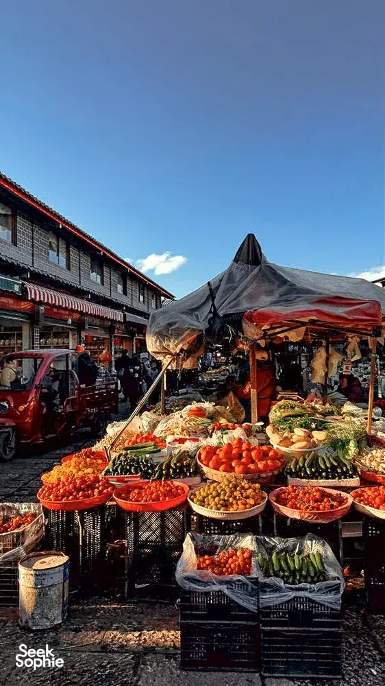 Open-air market in Lijiang Ancient Town piled high with fruit and vegetables in metal trays, under red awnings and blue sky