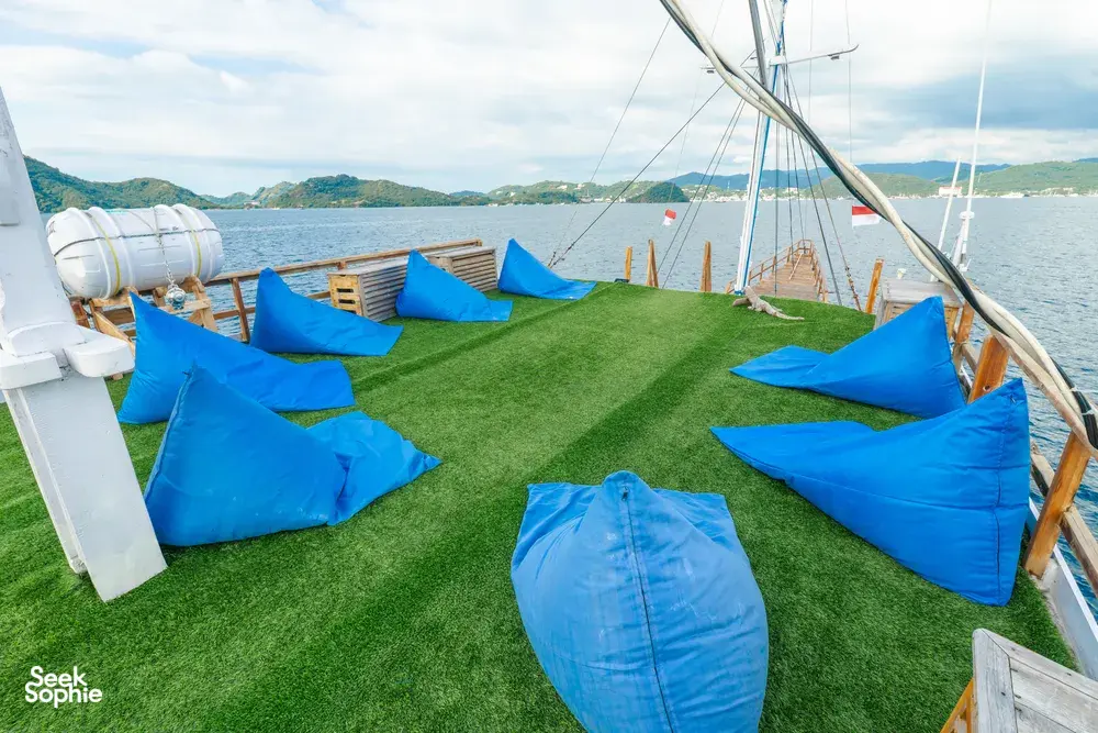 La Famille Komodo boat deck with green turf and blue beanbags laid out to relax, with sea and hills in the distance in Labuan Bajo