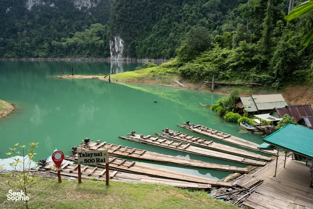 Old bamboo raft platforms and a floating jetty by a turquoise shoreline, framed by limestone hills and rainforest at Cheow Lan Lake