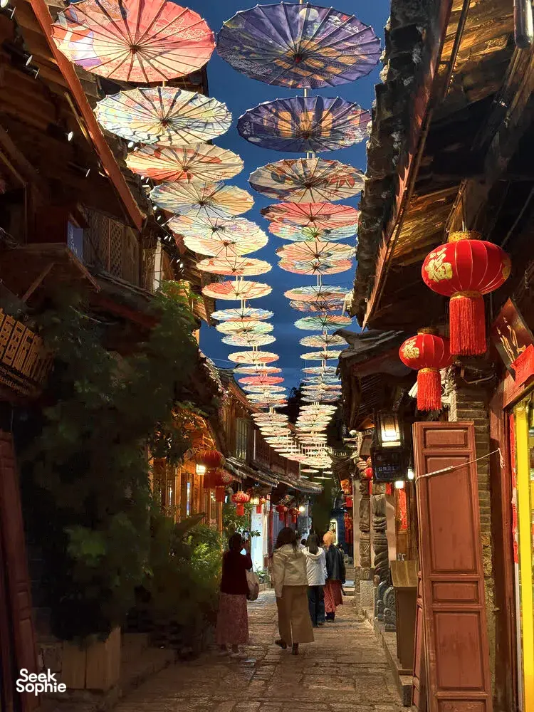 Lantern-lit lane in Lijiang’s old town with colourful paper umbrellas overhead as travellers stroll at dusk