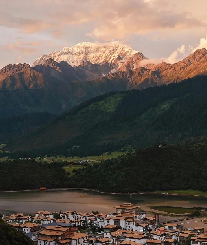 Snow-capped mountain range over a quiet valley town and reservoir during golden hour, framed by dark forested slopes