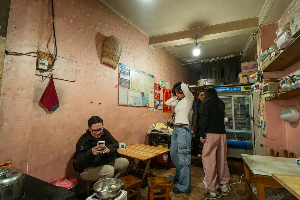 People in a small backstreet eatery kitchen, one seated with a phone while others stand preparing food