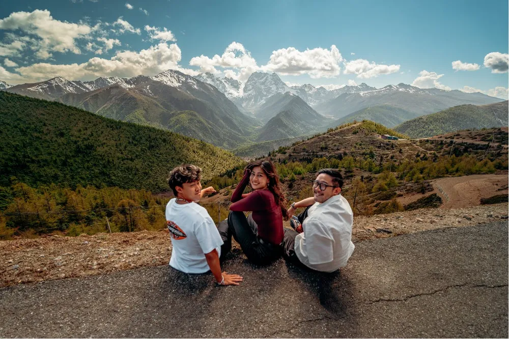 Travellers posing on a rocky mountain lookout with a wide valley of ridgelines and cloudy blue sky behind them
