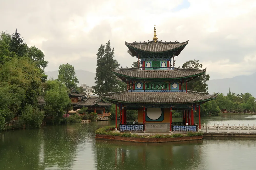 Traditional multi-tier pavilion on a lakeside platform, mirrored in calm water with greenery and distant hills under a cloudy sky