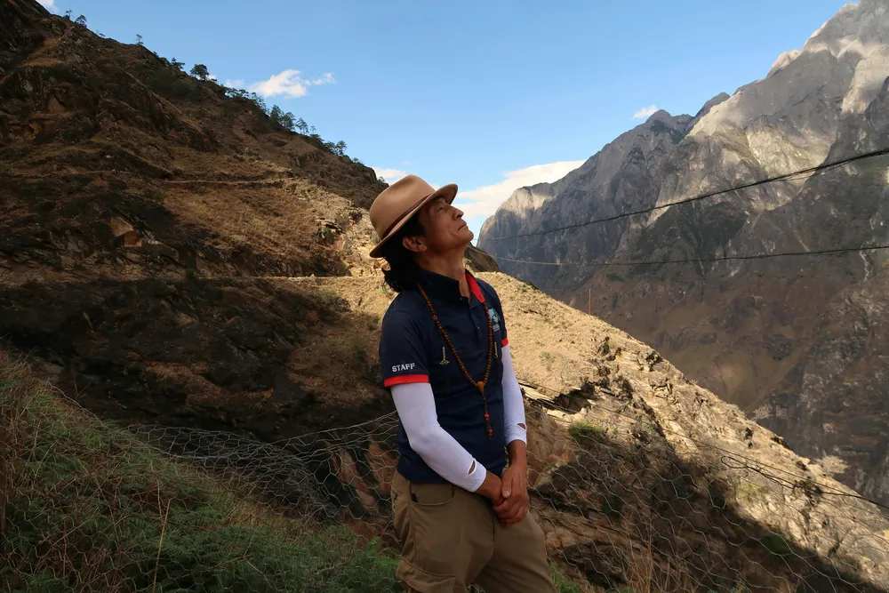 Hiker in a brimmed hat pausing on a rocky mountain trail, looking out over steep valleys and distant peaks under blue sky