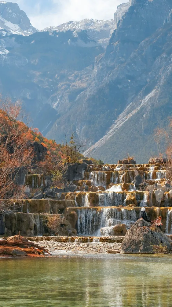 Cascading stone waterfall over terraced steps as blue mountains loom in the background, with calm water in the foreground