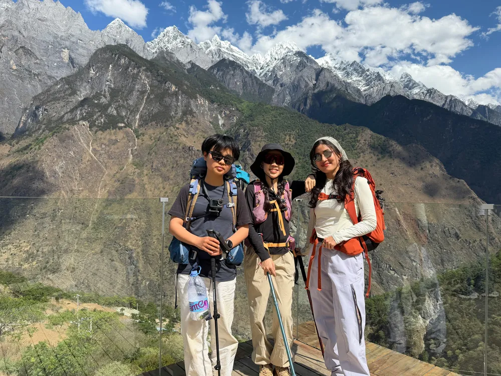 Three trekkers pose on a mountain overlook with wide-brim hats and backpacks, facing dramatic snowy peaks beneath clouds