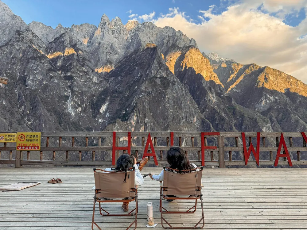 Travellers seated on a wooden deck overlooking rugged alpine peaks as the sun colours the cliffs in the distance