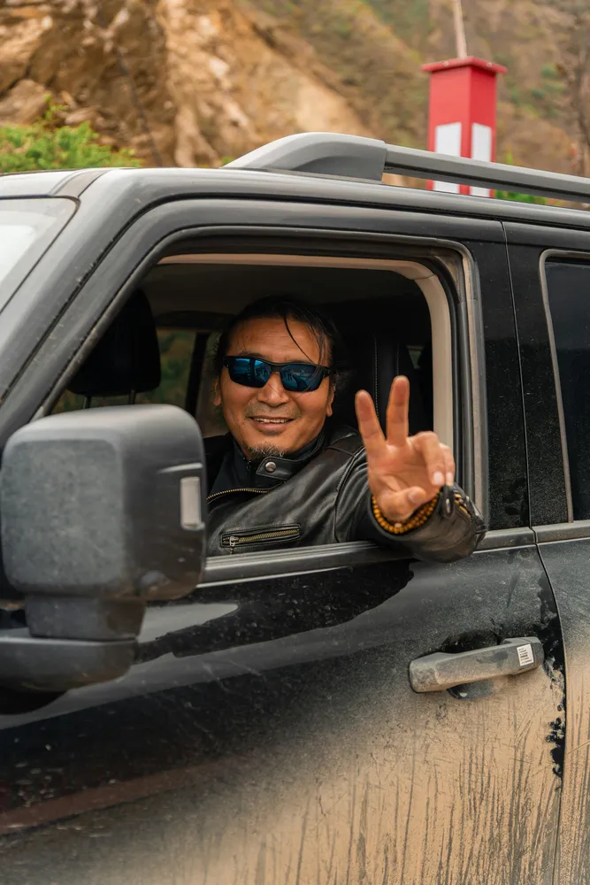 Driver smiling and flashing a peace sign from the open window of a rugged off-road vehicle on a winding mountain road