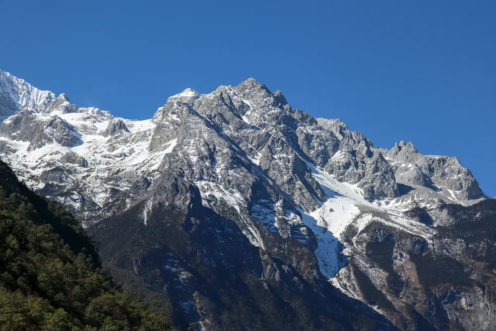 Puncak Gunung Salji Naga Jed dengan salji putih terang dan glasier di atas cerun dipenuhi pokok pain berhampiran Lijiang, Yunnan, di bawah langit biru yang cerah