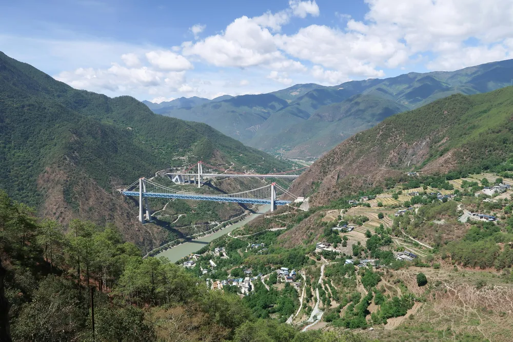 Suspension bridge crossing a river through a lush mountain gorge, with small villages below under billowing clouds