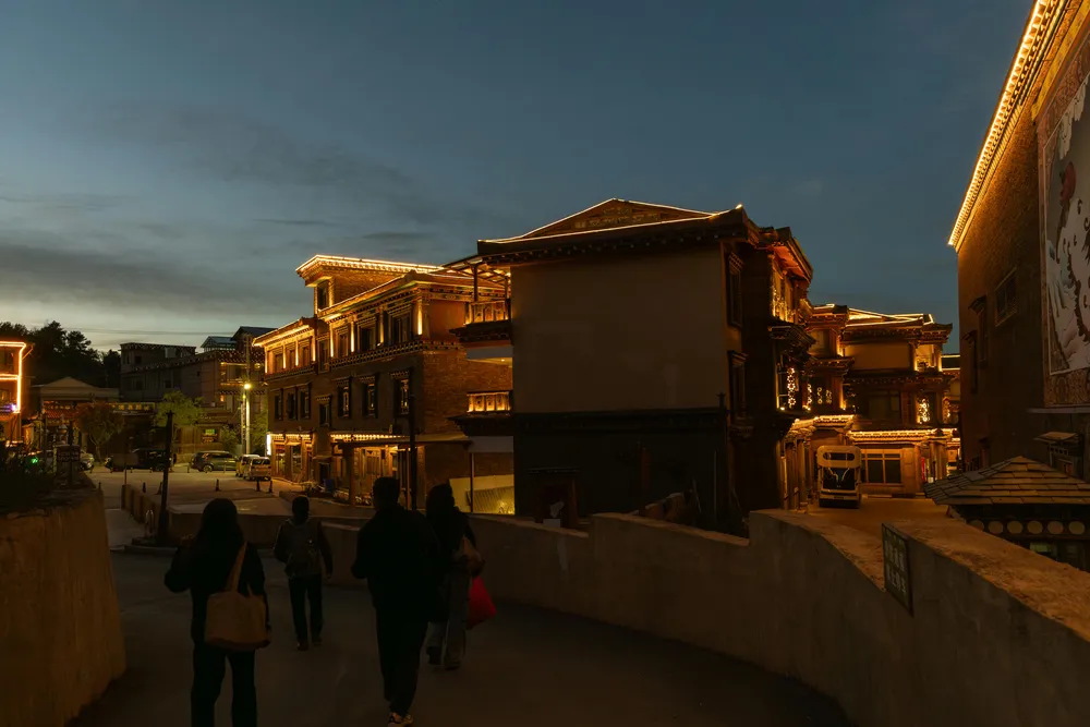 Evening street with warm lights along historic buildings, pedestrians walking past shopfronts under a twilight sky