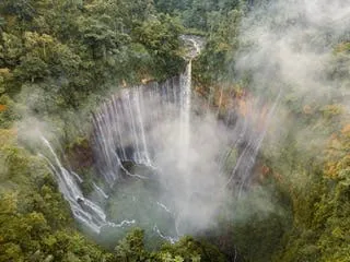 Aerial view of a wide waterfall plunging through dense rainforest canopy, sending mist across the gorge below
