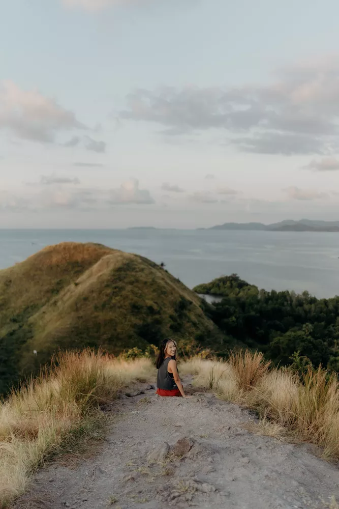 Woman sitting on a rocky trail atop grassy hills, overlooking the sea at dusk on a viewpoint in Labuan Bajo