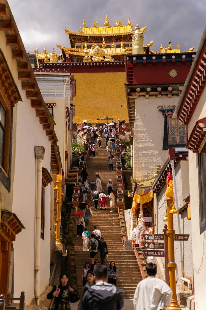 Visitors walking through a narrow corridor in an ornate palace complex, with golden rooflines, steps, and hanging lamps