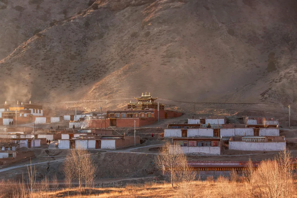 Tibetan-style monastery buildings clustered on a hillside beneath dramatic mountains and dusty evening light