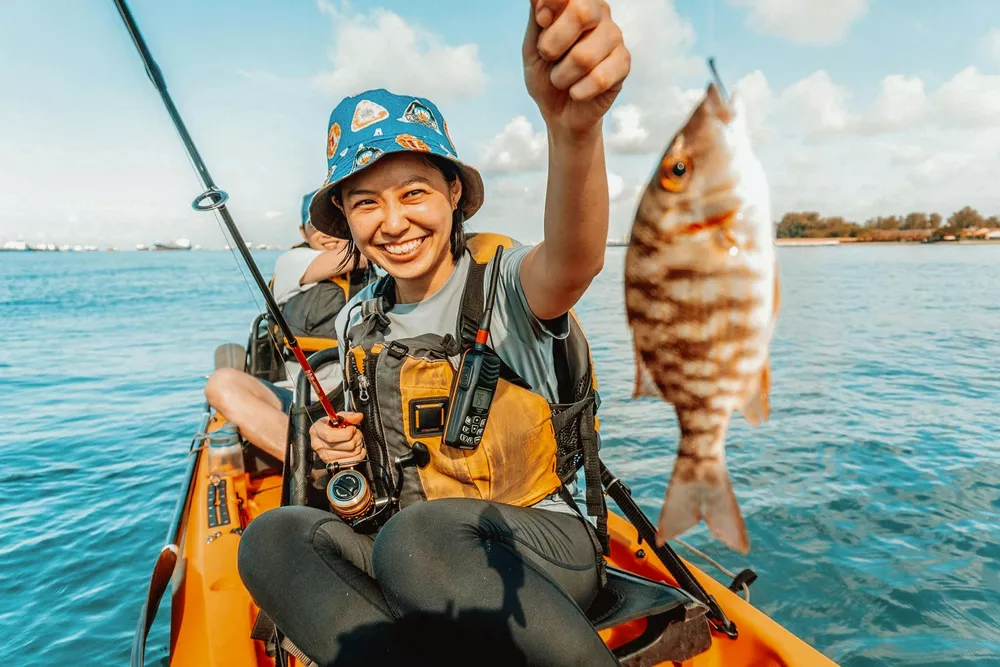Smiling kayaker on an orange inflatable boat holds up a caught fish, with fishing gear visible on the open water