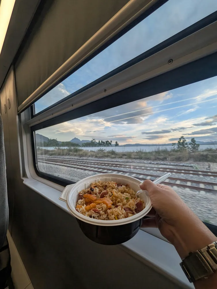 Hand holding a cup of noodles near a train window as tracks glide past the coast at sunset, with sea and distant hills outside
