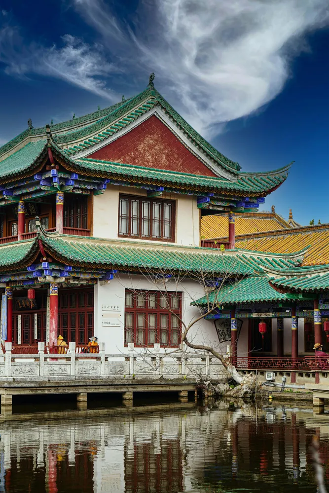Traditional palace building with layered green tiled roofs beside a pond, reflected in the water under a blue sky
