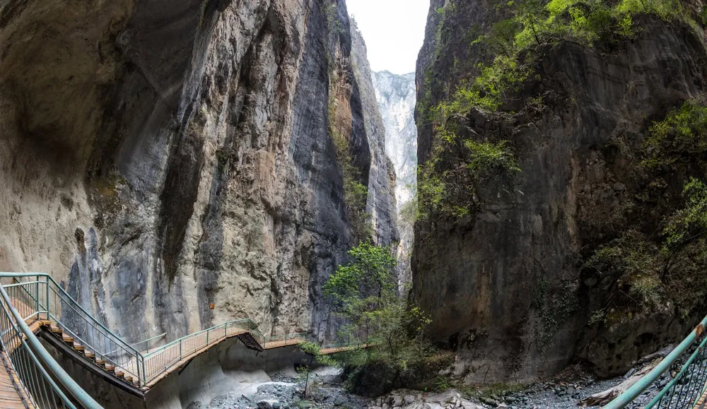 Steep rocky canyon with metal stairways descending into a narrow riverbed, framed by green trees on the cliff edges