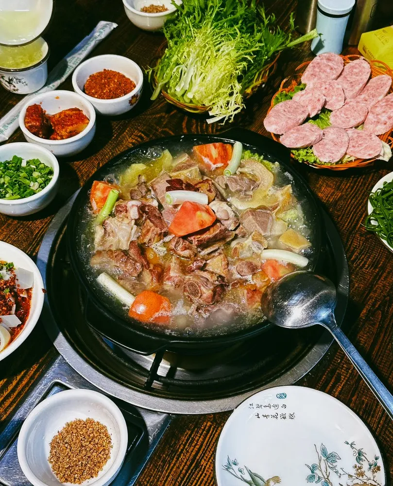 Hot pot meal on a table with a bubbling broth pot, sliced meats, leafy greens, and several dipping sauce bowls