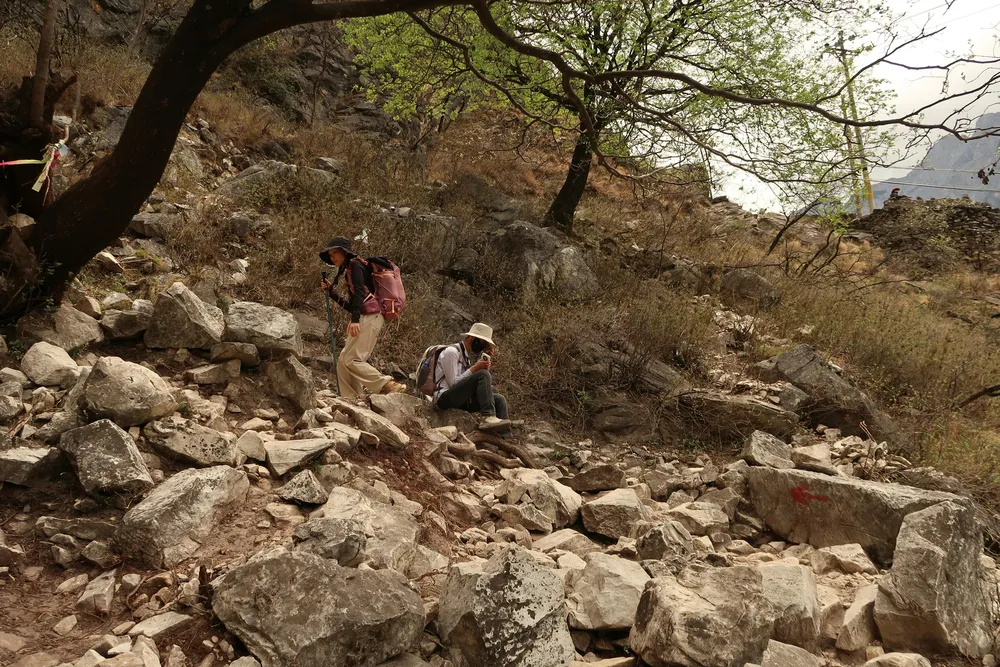 Backpackers rest on large rocks while a trekking guide talks, on a dry mountain hillside with scattered trees and a valley view