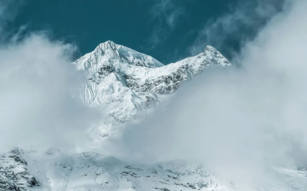 Snowy mountain peaks emerging from low clouds, with a sharp ridge and bright ice under a cold misty winter sky