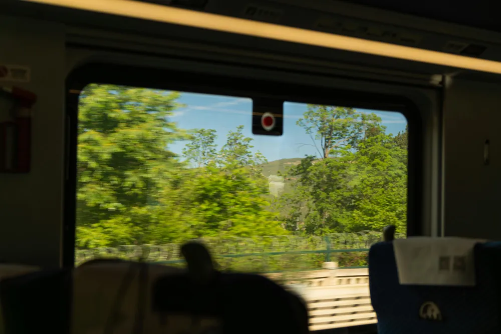 View from inside a vehicle through a curved window onto lush green hills and trees, with seated silhouettes in the foreground
