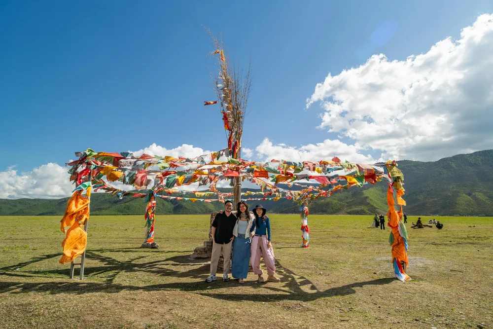 Travellers standing beside colourful prayer flags on a tall pole in a wide grassy valley with mountains under a bright blue sky