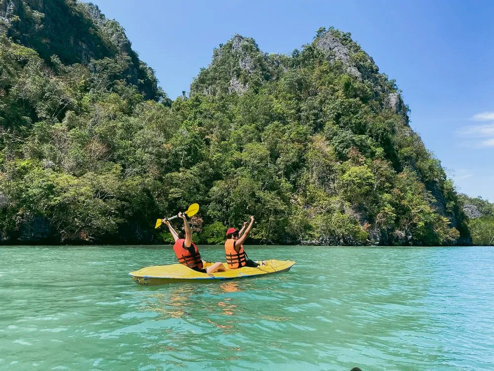 Kayakers in life jackets paddle across clear turquoise water beside lush limestone cliffs in Langkawi, Malaysia