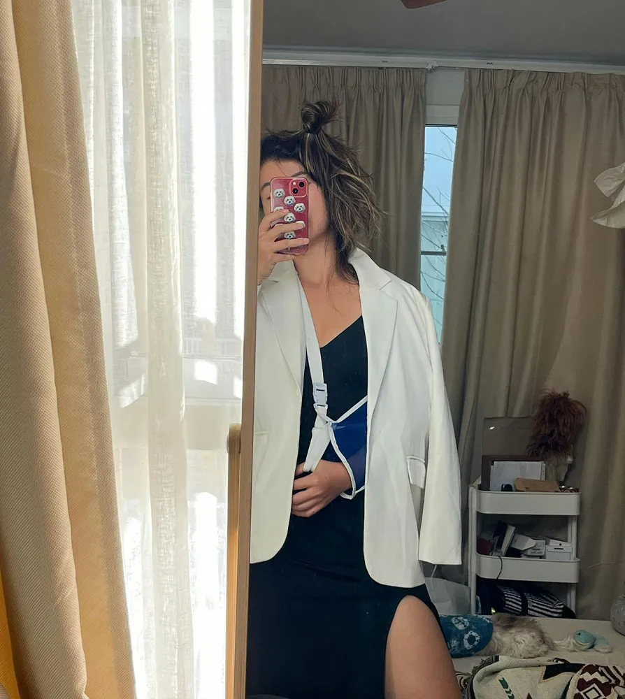 Person taking a mirror selfie indoors, wearing a white blazer over a dark dress while holding a smartphone