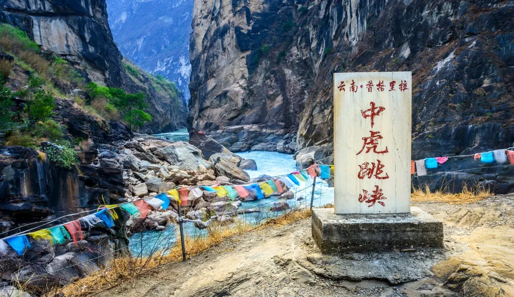 Rocky mountain gorge with a shallow river and sand banks, colourful prayer flags along the rocks and a stone sign bearing text