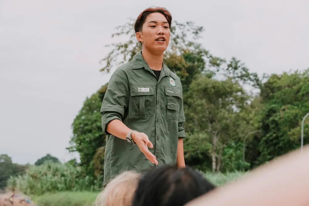Pemandu alam berpakaian seragam hijau bercakap kepada pengunjung di kawasan luar berhampiran pepohon yang rimbun, sambil memberi isyarat dan mereka memerhati dari dekat.