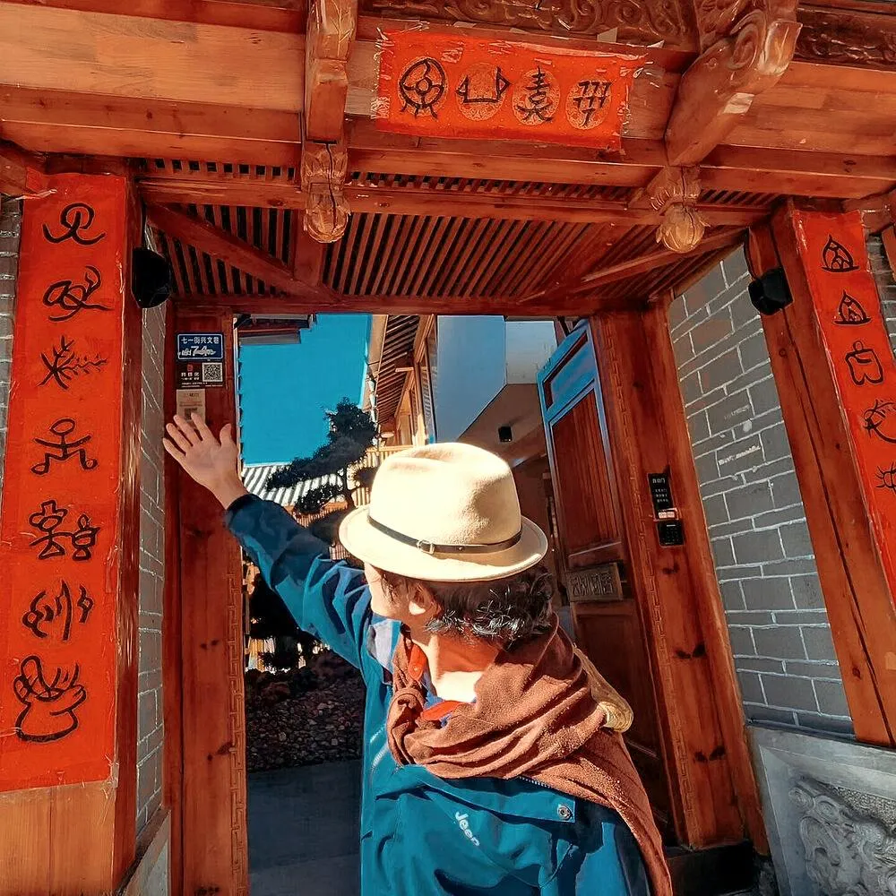 Person in a wide felt hat reaching for a wooden door with red calligraphy couplets, with a quiet courtyard beyond
