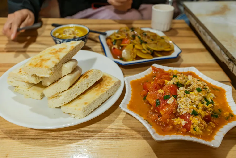 Plates of flatbread and mixed curry dishes on a wooden table, with diners’ hands and cups in the background