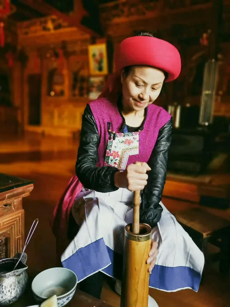 Cook wearing a pink hat and patterned apron stirs and grinds in a wooden mortar at a rustic kitchen counter, warm lantern light