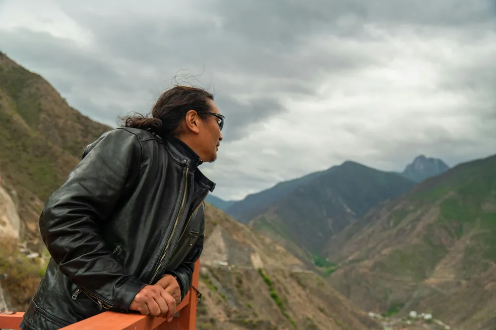 A hiker in a dark jacket gripping a wooden railing, looking out over misty mountain valleys beneath heavy clouds