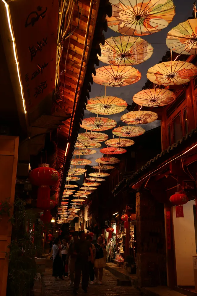 Lantern-filled narrow street with dozens of round paper umbrellas overhead, warm lights glowing as people walk between shopfronts