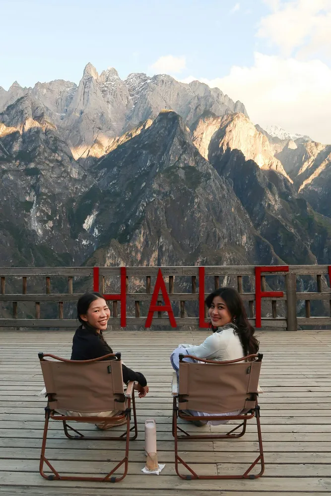 Two people sit in folding camp chairs on a wooden viewpoint, with dramatic cliffs behind them and red 'TRAIL' letters on rail