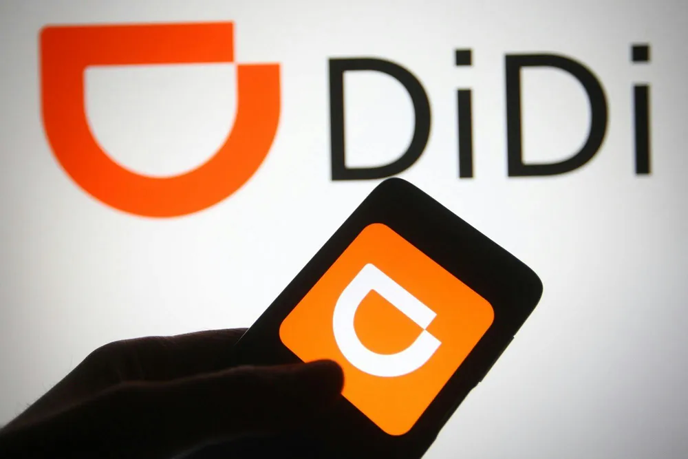 Hand holding a phone showing the DiDi app logo with the orange symbol and “DiDi” lettering on a light background