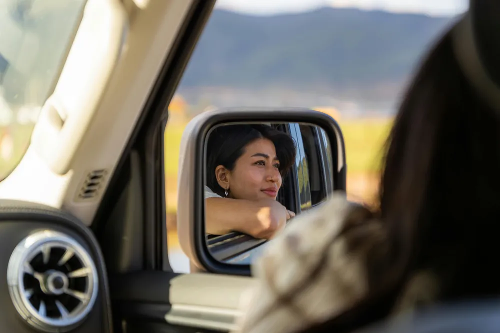 Woman smiling from the passenger seat, seen through a car side mirror, with mountains blurred outside the window