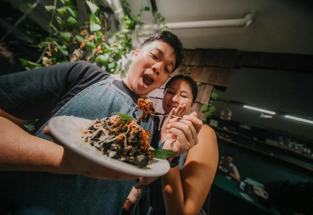 Man and woman smiling while sharing appetisers from a large takeaway plate in a restaurant kitchen with greenery backdrop