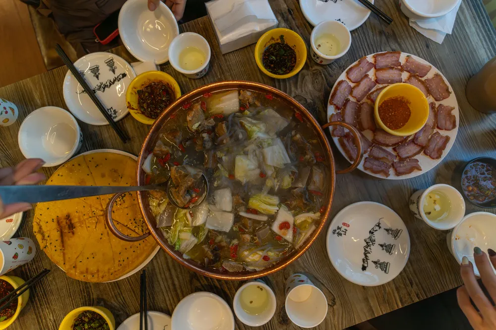 Overhead spread of a hotpot with vegetables and broth, alongside plates of cured meat and dipping sauces on the wooden table