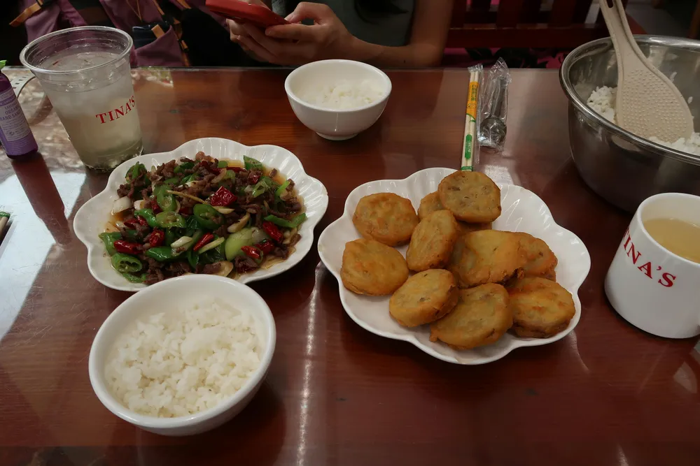 Wooden table spread with bowls of rice, a mixed vegetable salad and fried snacks, with drinks and cups at a casual meal