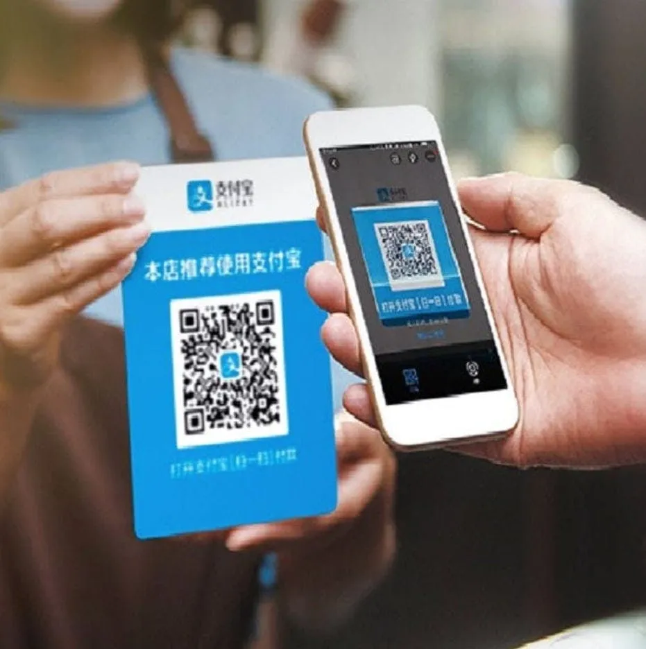 Hands holding a blue payment card and a phone showing a QR code for contactless mobile payment during travel