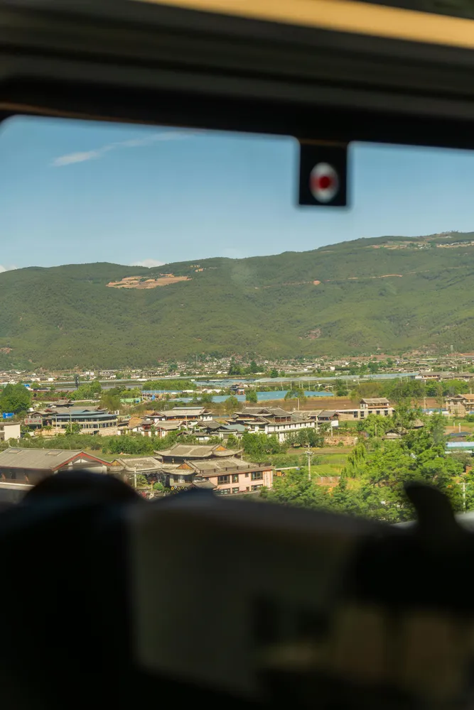 View of a hillside town and valley from inside a vehicle window, with green mountains and a small flag decal on the glass