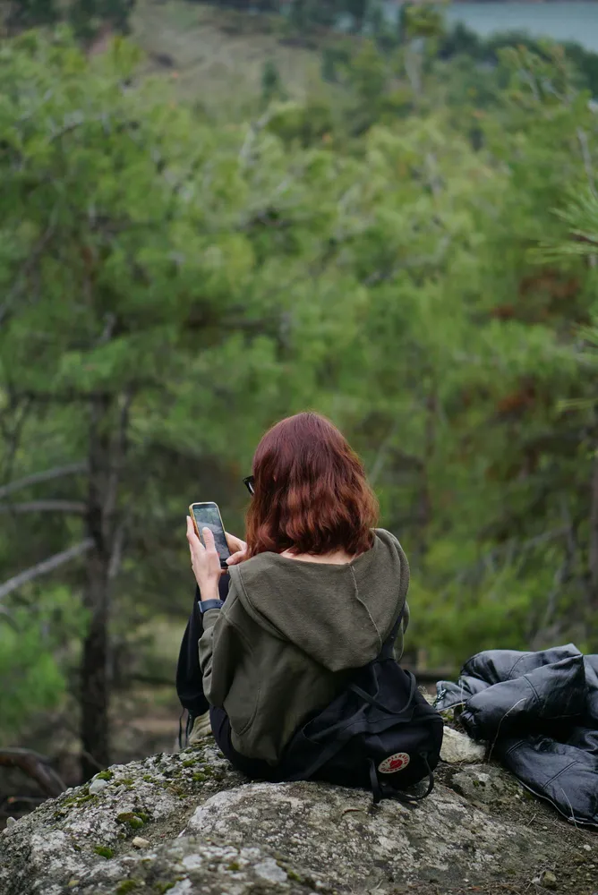 Back view of a hiker sitting on a mossy boulder with a phone, gazing over lush green forest slopes from a lookout